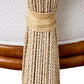 Close-up of a Strings Attached Lounge Chair with beige woven fabric and decorative long tassels tied with a ribbon on a natural-finish rattan frame.