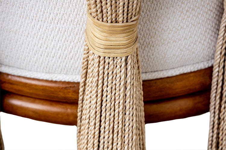 Close-up of a Strings Attached Lounge Chair with beige woven fabric and decorative long tassels tied with a ribbon on a natural-finish rattan frame.