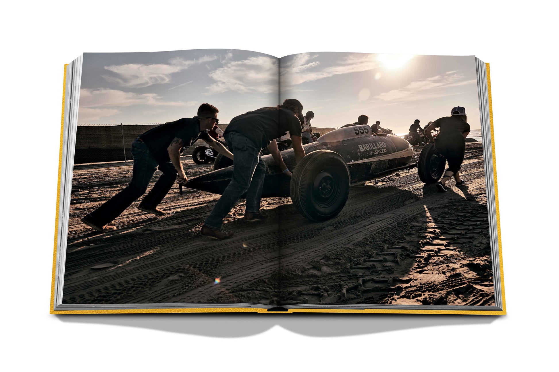 A team of individuals pushing a vintage race car on a sandy surface, captured in a photograph within an open book at The Race of Gentlemen by Assouline.