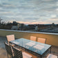 A rooftop terrace features six Janus et Cie Amalfi Dining Side Chairs in Slate around a glass-top table with stainless steel frames, all set against city buildings under a cloudy evening sky.