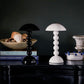 A black and a white Addison Ross Bobbin LED Cordless Lamp with spherical bases sit on a dark table alongside books, decor, and a bowl of blue beads—creating an elegant lighting scene against a moody backdrop.