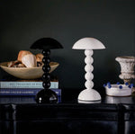 A black and a white Addison Ross Bobbin LED Cordless Lamp with spherical bases sit on a dark table alongside books, decor, and a bowl of blue beads—creating an elegant lighting scene against a moody backdrop.