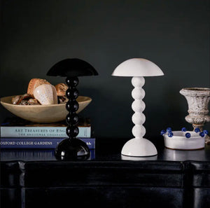 A black and a white Addison Ross Bobbin LED Cordless Lamp with spherical bases sit on a dark table alongside books, decor, and a bowl of blue beads—creating an elegant lighting scene against a moody backdrop.