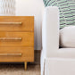 Close-up of a light-colored sofa positioned next to the Ascue Nightstand. The nightstand features three drawers with distinct rattan-wrapped pulls, enhancing its Mid-century design. A lamp sits atop the nightstand, while a green-striped pillow rests on the sofa.