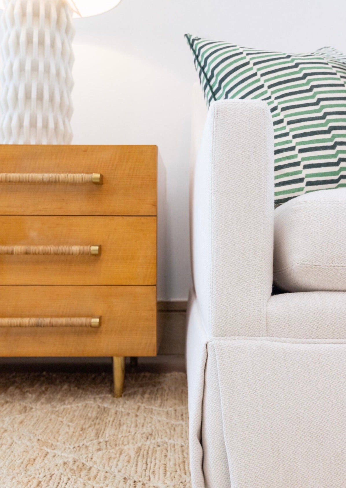 Close-up of a light-colored sofa positioned next to the Ascue Nightstand. The nightstand features three drawers with distinct rattan-wrapped pulls, enhancing its Mid-century design. A lamp sits atop the nightstand, while a green-striped pillow rests on the sofa.