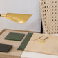 Close-up of a minimalist desk setup featuring the Pigeon and Poodle Asby Desk Set in Forest, including green notebooks, a magnifying glass, a gold and gray pen, and a gold desk lamp. A woven-back chair is partially visible in the background, complemented by accessories from this refined collection with versatile finishes.