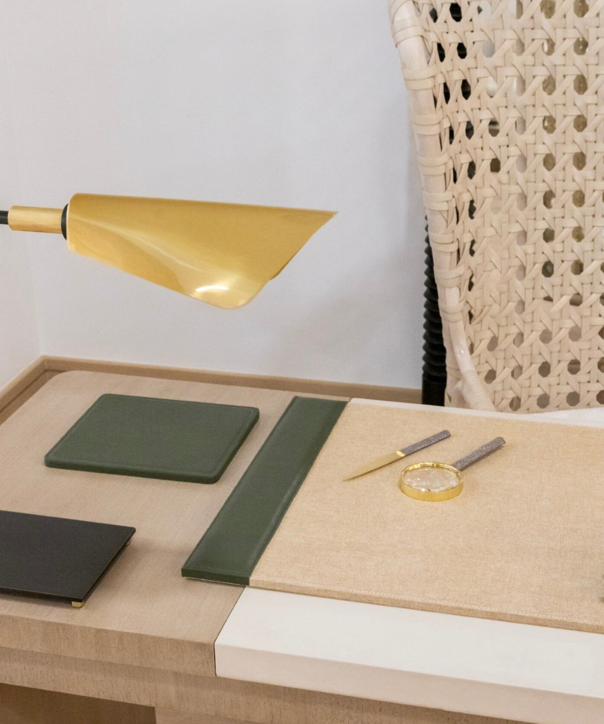 Close-up of a minimalist desk setup featuring the Pigeon and Poodle Asby Desk Set in Forest, including green notebooks, a magnifying glass, a gold and gray pen, and a gold desk lamp. A woven-back chair is partially visible in the background, complemented by accessories from this refined collection with versatile finishes.