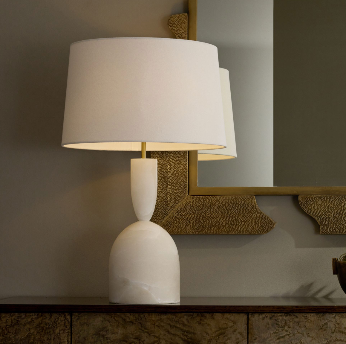 The Brighton Lamp, with its white curved base and cylindrical shade, featuring an antique brass three-way rotary switch, sits on a wooden surface next to a textured mirror.