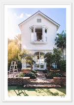 A white, two-story house with an open second-floor window. A ladder is placed on a brick-paved path in the garden, lined by a charming hedgerow and surrounded by plants and trees. Nick Mele Fine Art The Nest.