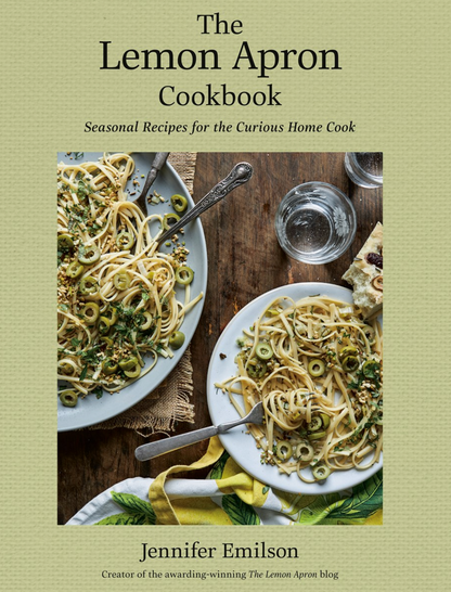 Cover of "Lemon Apron Cookbook" by Jennifer Emilson, featuring a photograph of two plates of spaghetti with herbs and olives, a glass of water, and bread on a wooden table. This beautiful cover perfectly captures the essence of rustic recipes and seasonal cooking.