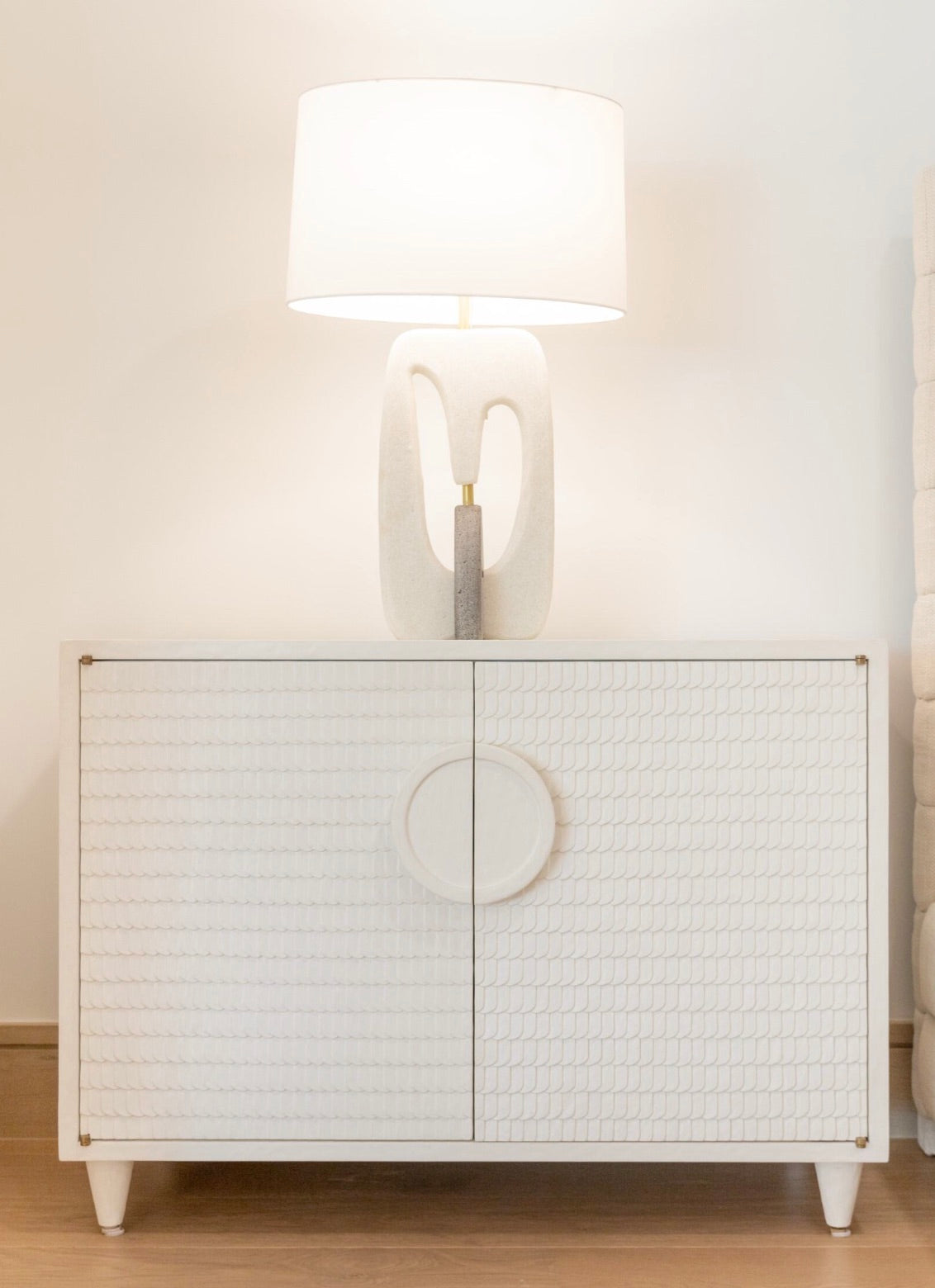 A white table lamp with a modern design sits atop the Lance Cabinet, a hand-crafted piece featuring a textured pattern, adding a minimalist accent to the room.