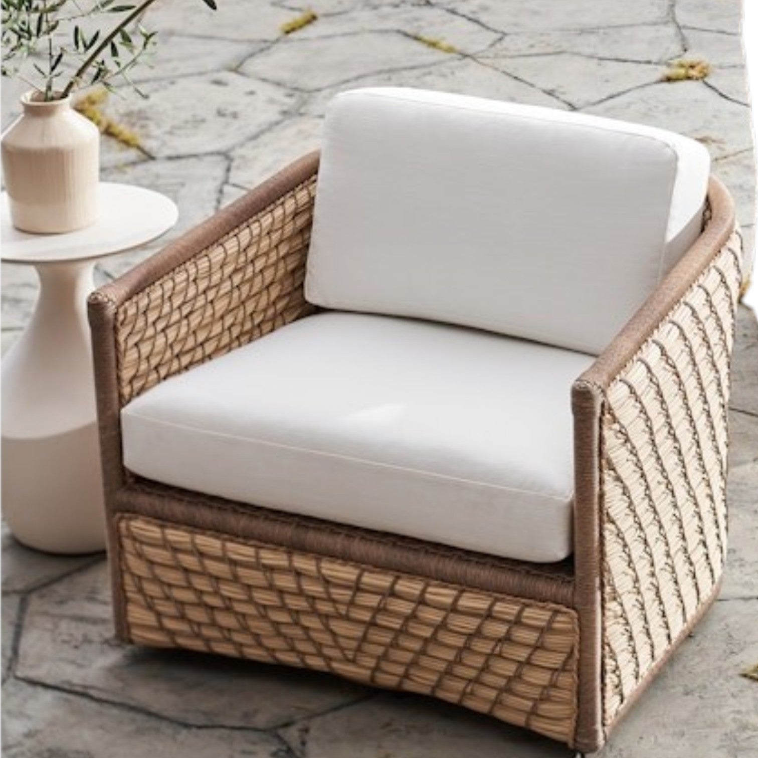 The Capitola Swivel Lounge Chair, featuring woven rattan and white cushions, sits by a small round side table on a stone patio; its powder-coated aluminum frame ensures outdoor durability.