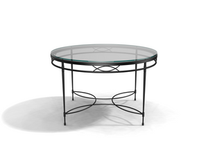 The Amalfi Glass Top Round Dining Table features a hand-forged steel frame with decorative curved metal accents on the legs and base, shown on a white surface.