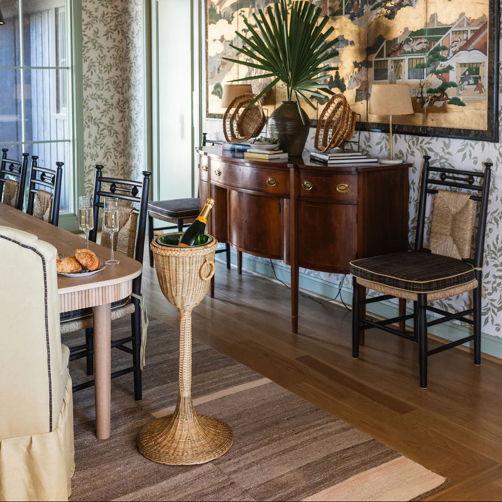 The Champagne Stand holds a bottle beside a dining table set with glasses and bread, with a sideboard, decor, and chairs visible in the background.