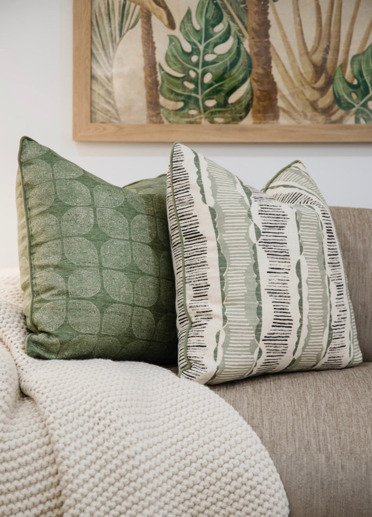 Two Petaluma Moss Pillows rest on a beige sofa with a knitted throw blanket, beneath a framed artwork featuring botanical designs. The attention to detail and use of the finest materials elevate the cozy setting.