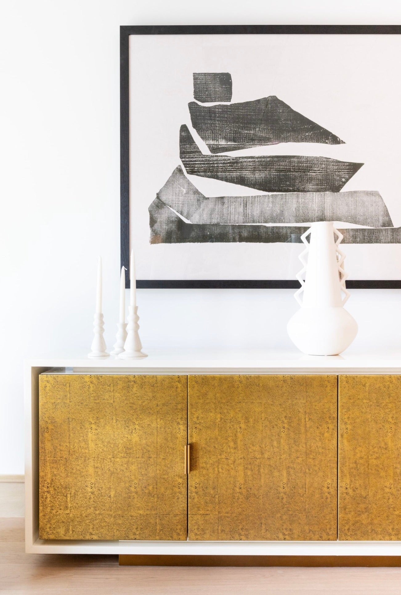 The Diego Four Door Cabinet, featuring high gloss ivory vellum doors, sits against a white wall. It is adorned with two white candlesticks, a white vase, and topped with a framed abstract black and white artwork.