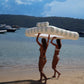 Two women in swimsuits carry a SUNNYLiFE Luxe Twin Hammock Float above their heads on a sandy beach near boats and forested hills.