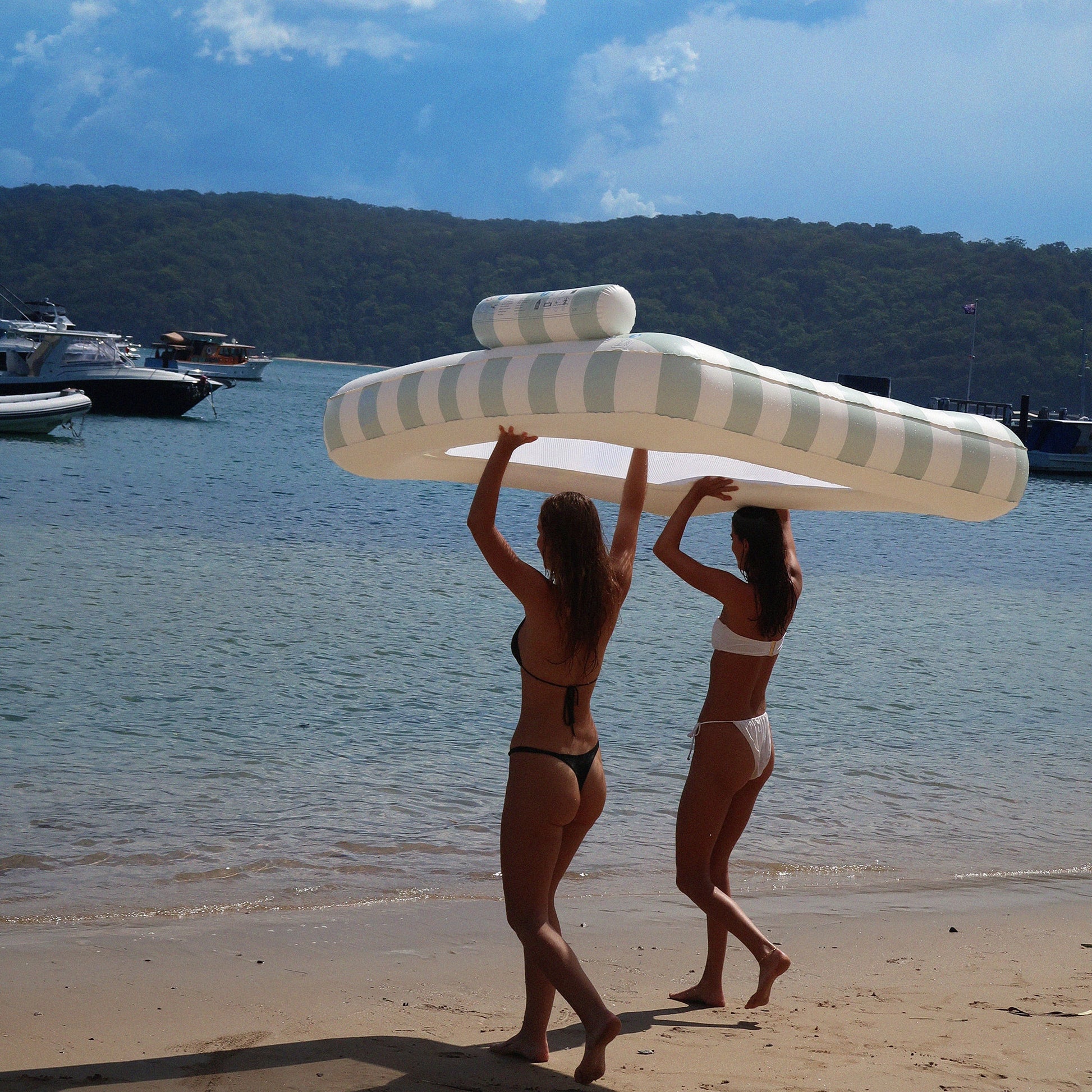 Two women in swimsuits carry a SUNNYLiFE Luxe Twin Hammock Float above their heads on a sandy beach near boats and forested hills.