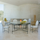 A modern dining area features a round glass table, four white upholstered chairs, the Amalfi Round Back Armchair, a bowl of apples, and a fireplace against a light, minimalist backdrop.