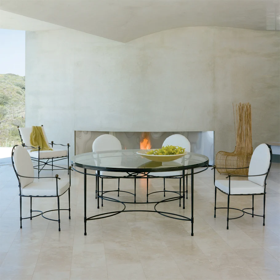 A modern dining area features a round glass table, four white upholstered chairs, the Amalfi Round Back Armchair, a bowl of apples, and a fireplace against a light, minimalist backdrop.