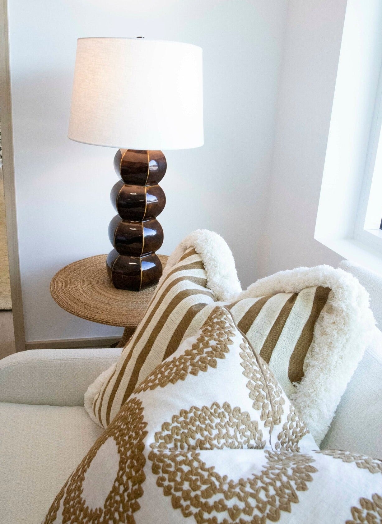 A modern lamp with a white shade sits on a round wicker table next to a cream sofa adorned with custom-made Alexander Epices Pillows featuring patterned brown and white cushions, showcasing special attention to detail.