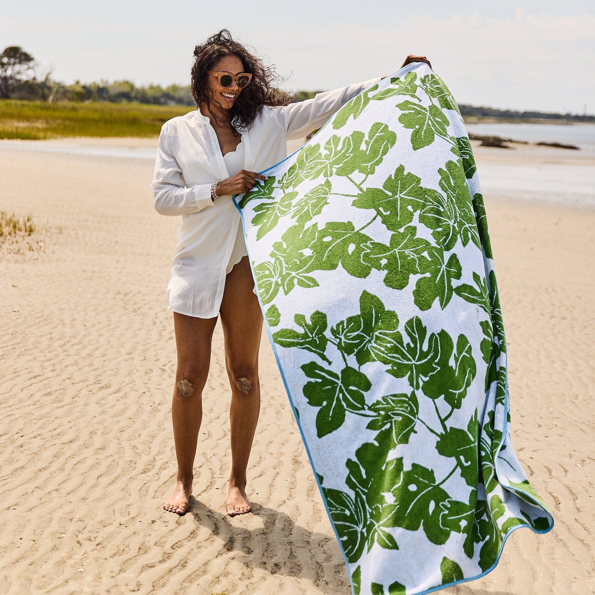 A woman stands on a sandy beach holding an oversized Weezie x Peter Dunham Fig Beach Towel adorned with green leaf patterns. She wears sunglasses and a white shirt, enjoying the perfect blend of style and comfort.