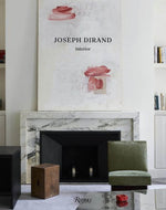 A minimalist living room featuring a fireplace with a marble surround, a green chair, a wooden block, and a large art piece titled "Joseph Dirand: Interior" above the mantle, embodying the elegance of French style.