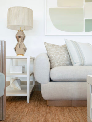 A beige modern living room features a light gray armchair adorned with Tangier Celadon Pillow Down, a white side table holding a geometric lamp and decor items, and soft-toned abstract wall art, all crafted with the finest materials.