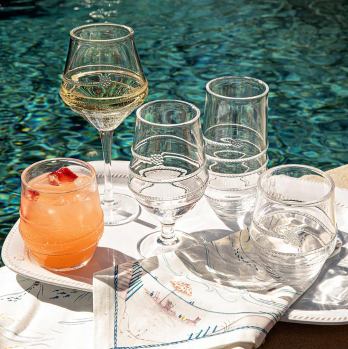 Tray featuring two empty wine glasses, two empty water glasses, and one glass filled with an orange drink from the Juliska Amalia Acrylic Collection, set on a towel beside a sunlit pool—ideal for the adventurous entertainer who appreciates practical indulgence with durable acrylic drinkware.