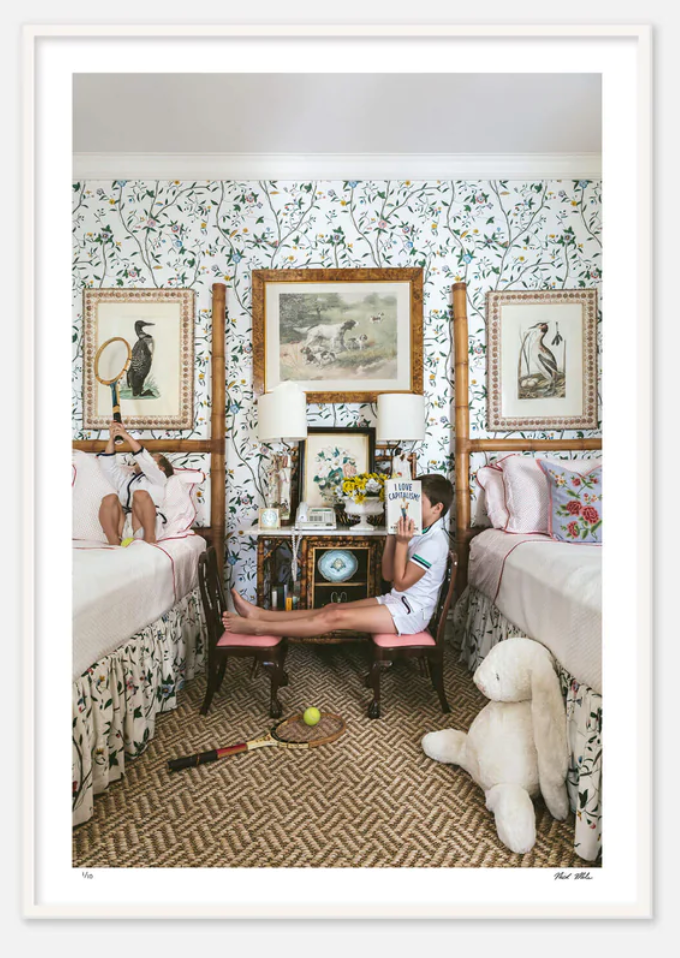 Two children are in a vintage-style bedroom with twin beds, reminiscent of Nick Mele Fine Art I Love Capitalism. One child is lying on a bed, hidden by pillows; the other sits on a chair between the beds, reading a book. Toys are scattered on the floor, creating one of those beautifully imagined scenes.