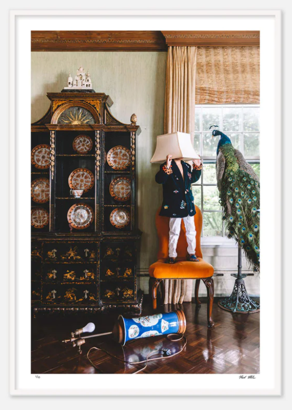 A young child stands on a chair in a living room, wearing a Nick Mele Fine Art Floor Lamp on their head. A China cabinet with ornate dishes and a peacock sculpture evoke gilded age glamour. An overturned blue canister is on the floor, capturing the whimsical essence of Newport RI as seen through Nick Mele's lens.