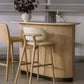 The Anton Bar, a small modern table, features two light wooden chairs, a vase with dark leaves, a metal cocktail shaker, and two crystal glasses atop its honey marble surface.