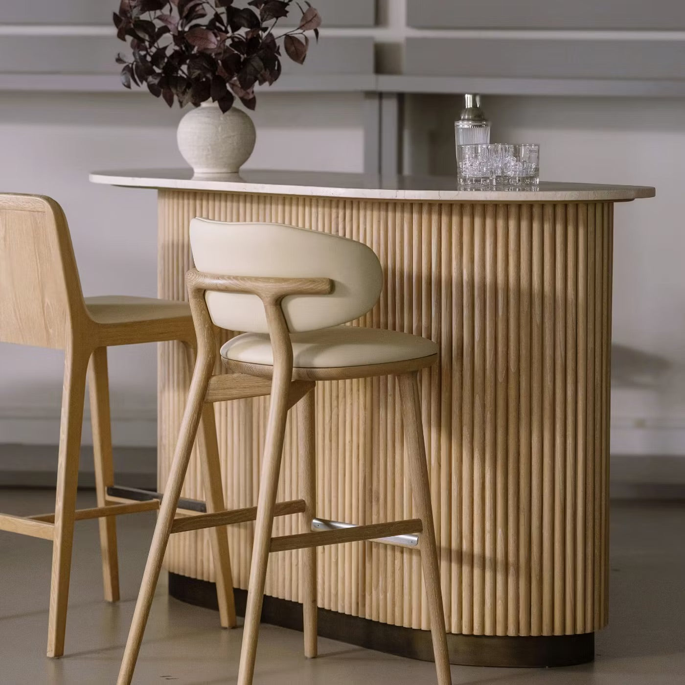The Anton Bar, a small modern table, features two light wooden chairs, a vase with dark leaves, a metal cocktail shaker, and two crystal glasses atop its honey marble surface.