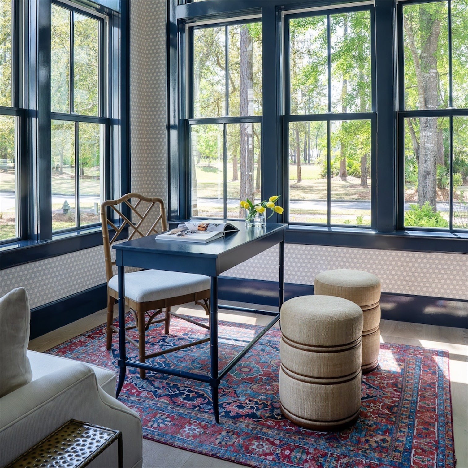 A bright home office with large windows, a black desk with papers and yellow flowers, a wooden chair, two Quinn Stools in Natural/Saddle, and a patterned rug on the floor.