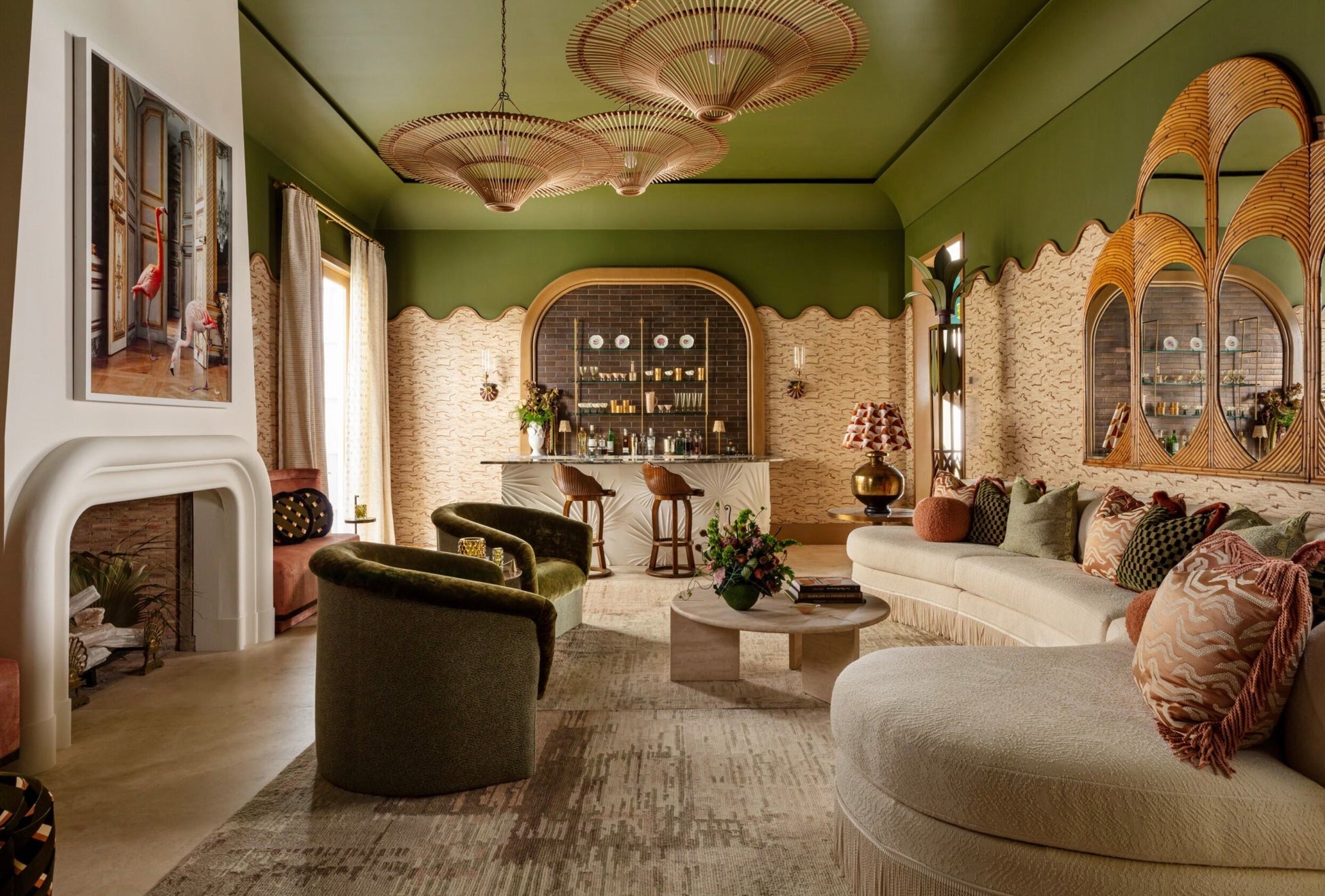 A stylish living room with green walls, beige and green furniture, a bar area, large mirrors, unique ceiling lights, and a decorative fireplace featuring a modern painting on the wall. A Lorenzo Cocktail Table adds an elegant touch while a sculptural piece completes the sophisticated ambiance.