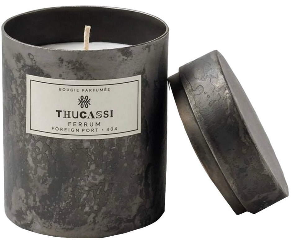 A scented candle in a matte dark container labeled "Thucassi Ferrum Candle Collection, Foreign Port" with the lid resting beside it, exudes a sweetly complex cardamom aroma.