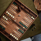 A hand-crafted wooden backgammon board with green and white checkers is set up for play. Two dice show the numbers 1 and 5. A brown dice cup and additional dice are placed nearby, showcasing the elegance of this natural wood Matis Backgammon Set.