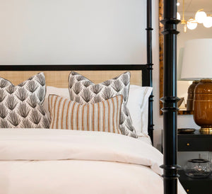 A neatly made bed featuring Palm Parade Graphite Pillows and white bedding, showcasing special attention to detail, next to a nightstand with a brown lamp and a glass container.