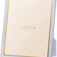 The AERIN Classic Piero Leather Frame Collection features a white standing picture frame with gold detailing and the word "AERIN" in the center, offering an elegant display fit for any room.