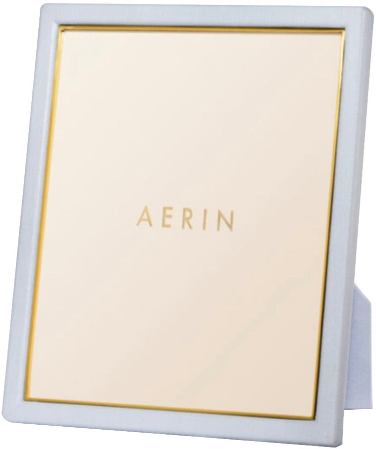 The AERIN Classic Piero Leather Frame Collection features a white standing picture frame with gold detailing and the word "AERIN" in the center, offering an elegant display fit for any room.