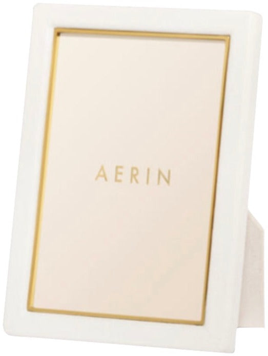 The AERIN Classic Piero Leather Frame Collection in white, featuring gold detailing and a beige background with the word "AERIN," creates an elegant display.