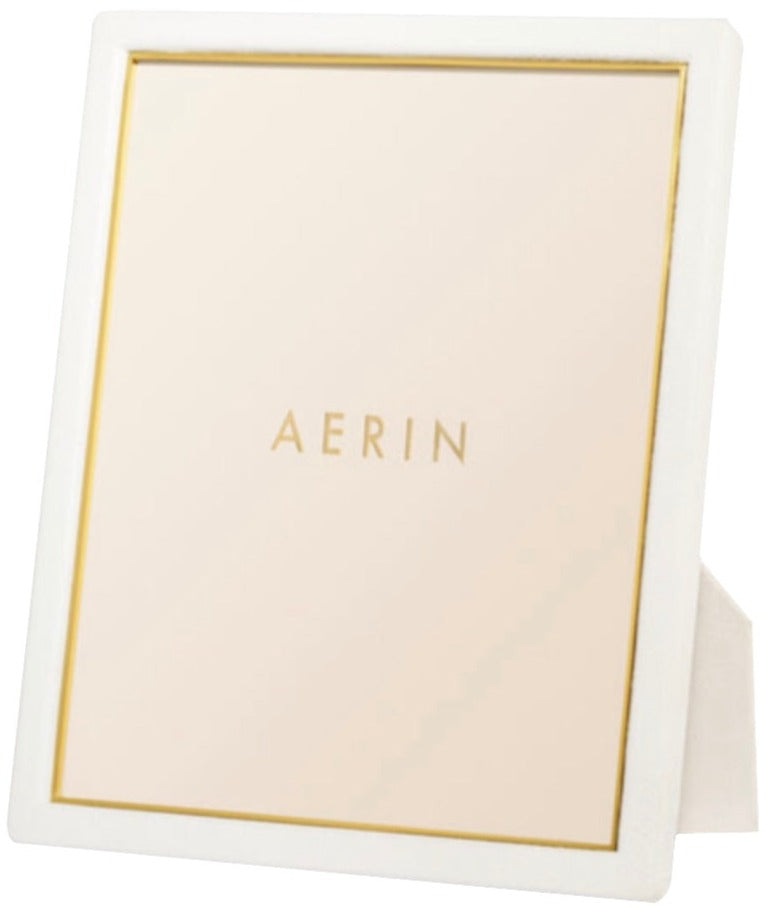 A tabletop mirror from the AERIN Classic Piero Leather Frame Collection, featuring the word "AERIN" in gold lettering centered on its reflective surface and framed by a white border with slim gold detailing, creates an elegant display.