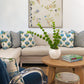 A living room with a beige sectional sofa, blue and white patterned pillows, a round wooden coffee table with a white vase and books, and floral wall art featuring Shell 2 Art by Paule Marrot from the Art Deco collection.