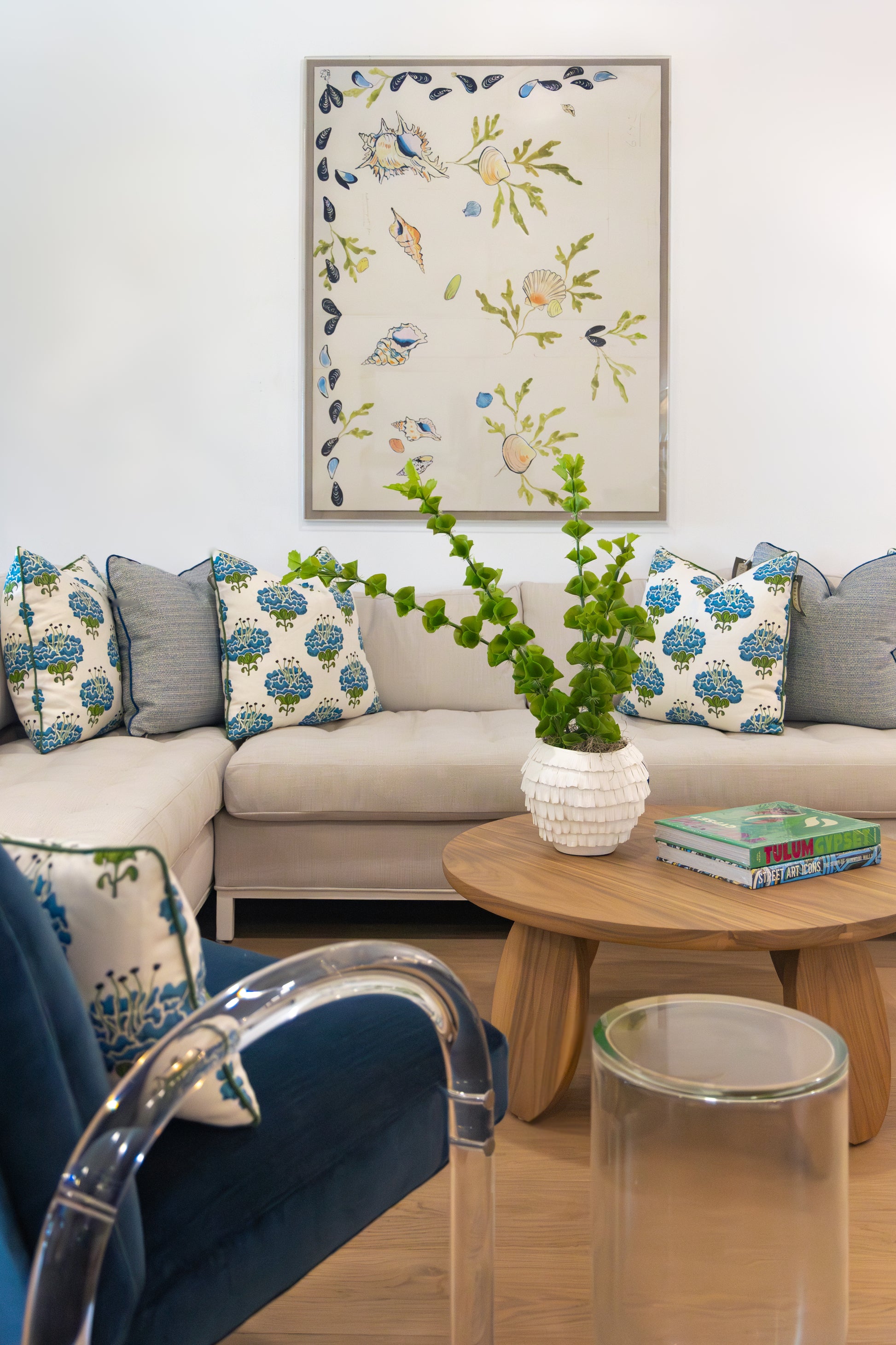 A living room with a beige sectional sofa, blue and white patterned pillows, a round wooden coffee table with a white vase and books, and floral wall art featuring Shell 2 Art by Paule Marrot from the Art Deco collection.