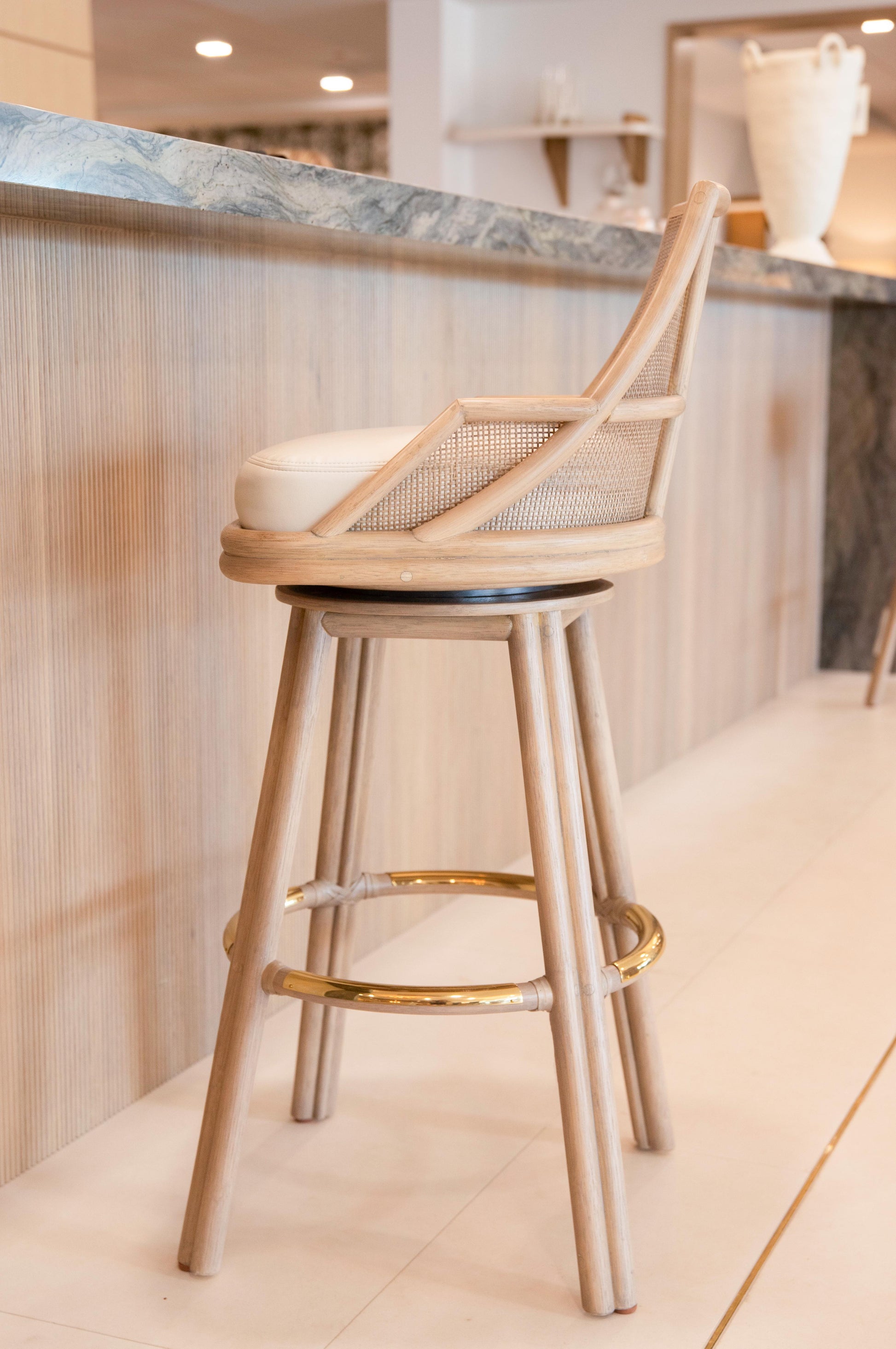 The Hayes Barstool, featuring a rattan frame and a caned interior backrest, boasts a white cushion seat and a gold footrest. This elegant stool is perfectly situated in front of a wooden counter with a marble top.