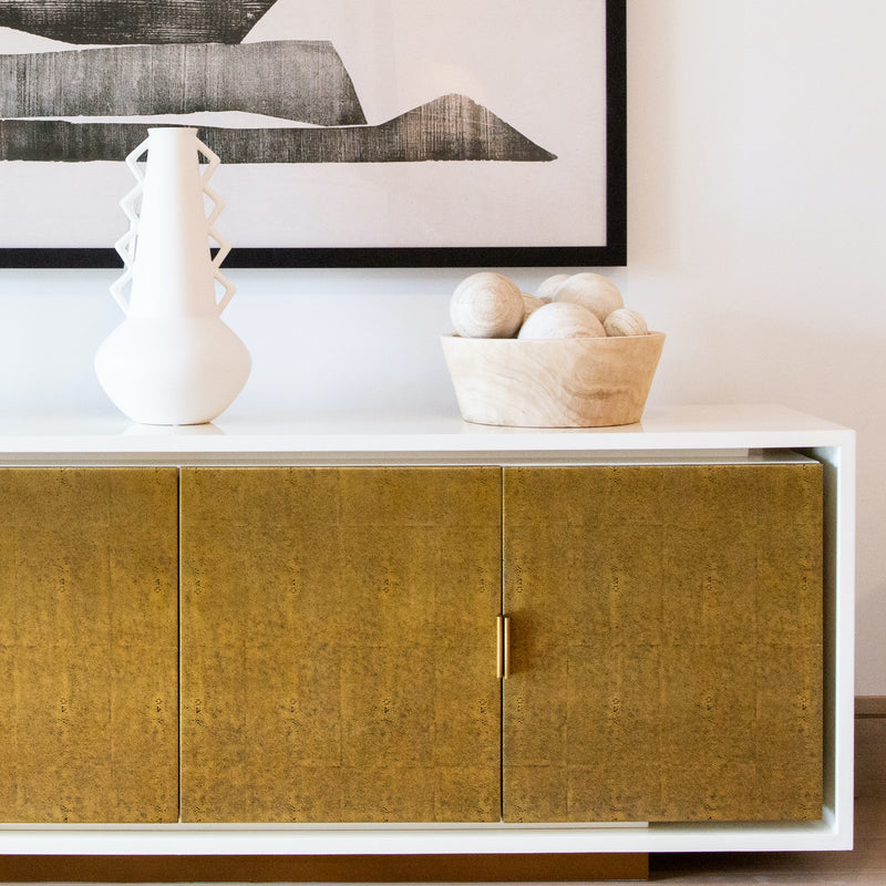 sideboard with gold metal doors, a white angular vase, decorative spheres in a bowl, and abstract black and white art. 