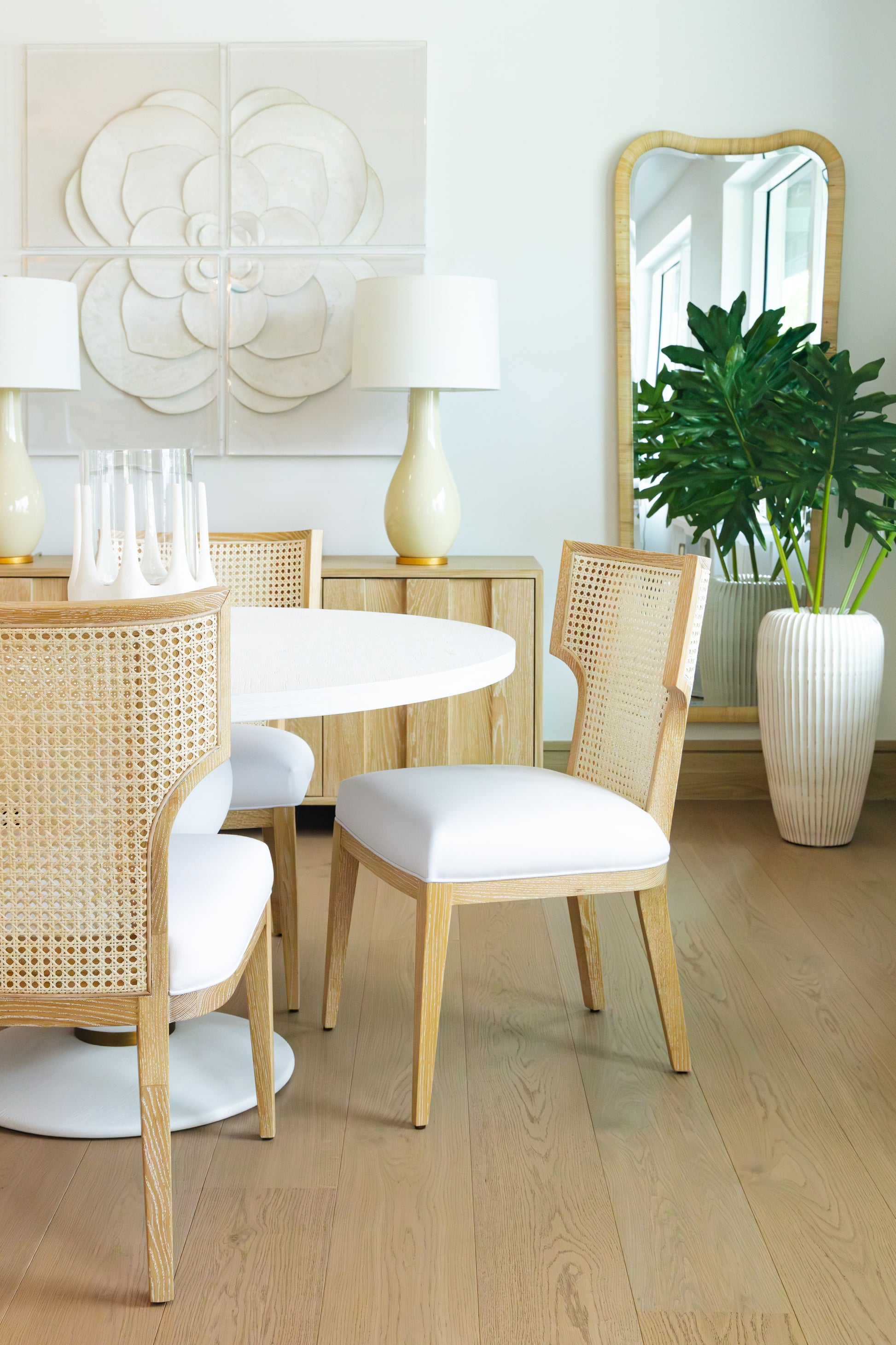 A modern dining area featuring rattan chairs, a round white table, a wooden sideboard with two lamps, a large green plant in a white pot, and the eye-catching Anela Mirror on the wall.