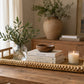 The Kenis Braided Rectangle Tray sits on a wooden table, holding a glass vase with greenery, stacked books, a wooden bowl, and a lit candle; a large vase and picture frame are in the background.