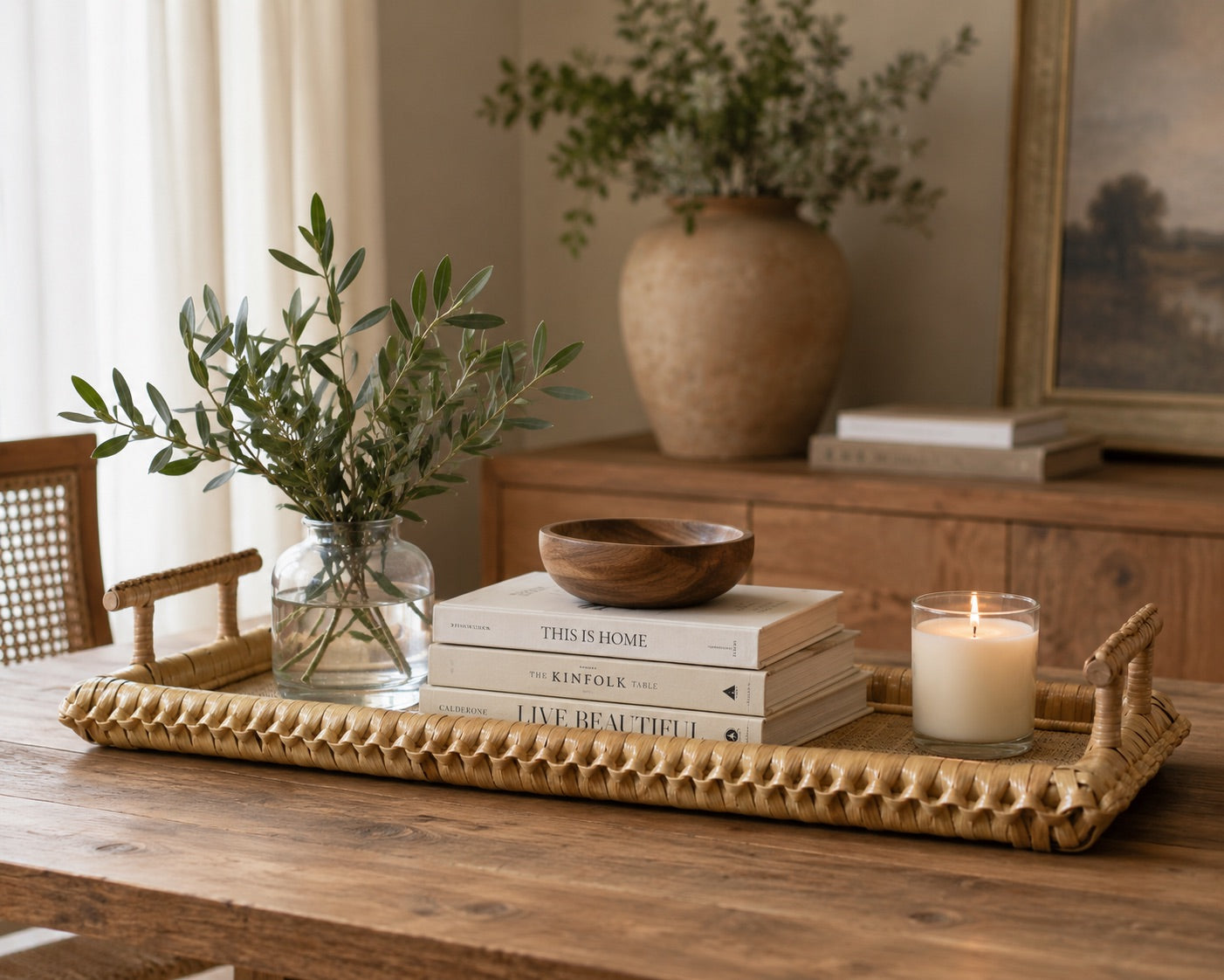The Kenis Braided Rectangle Tray sits on a wooden table, holding a glass vase with greenery, stacked books, a wooden bowl, and a lit candle; a large vase and picture frame are in the background.