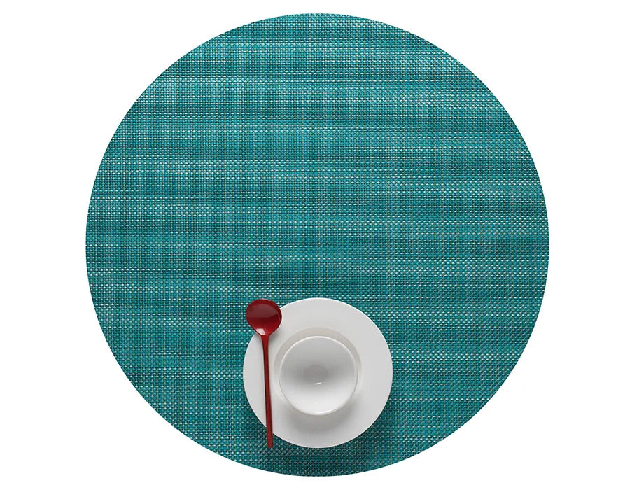 A Mini Basketweave Round Placemat in turquoise holds a white cup and saucer with a red spoon on top, viewed from above—ideal for bringing vibrant color to your dining table.